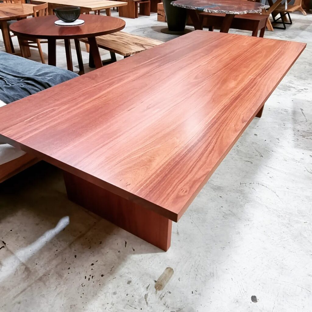 A large rectangular dining table with a smooth, polished jarrah timber top featuring warm reddish-brown tones and natural wood grain, supported by solid slab legs, displayed in a furniture workshop.